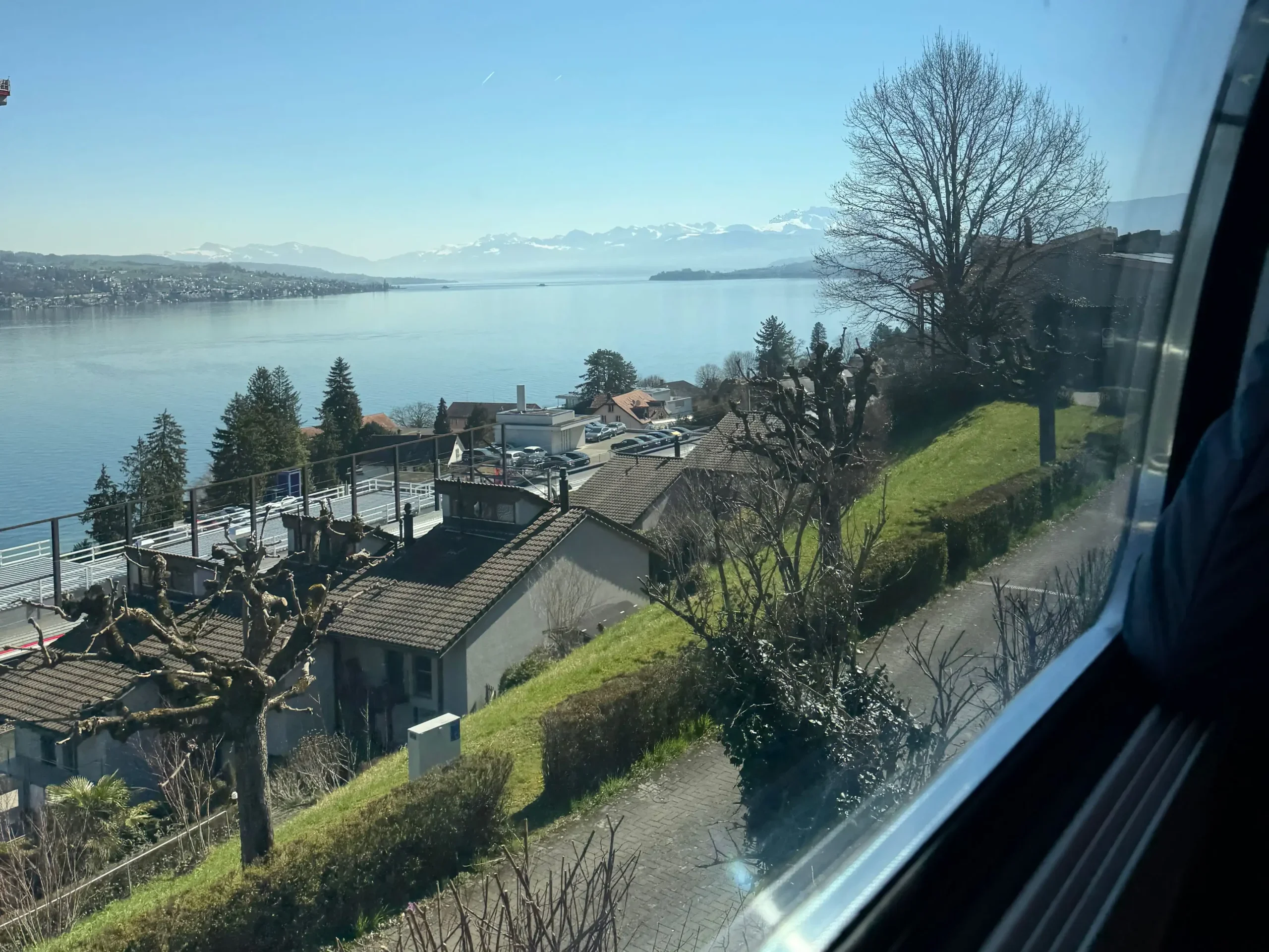 Tren camından süzülen manzara: göl kıyısında sessiz evler, uzakta karla örtülü zirveler.